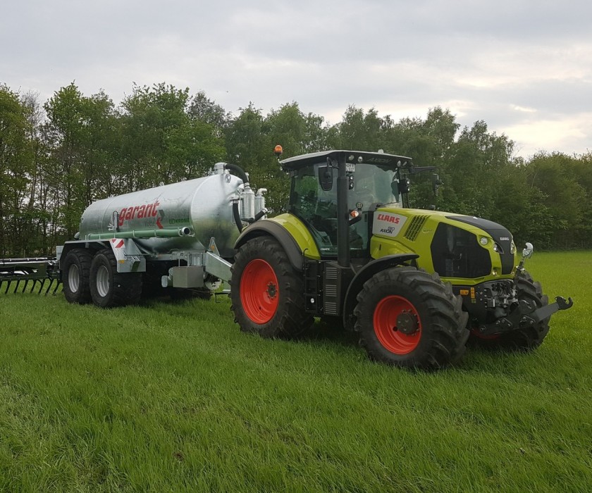 Grüner Claas Traktor mit anhängendem grauen Güllefass von Garant. Stehend auf einem Feld mit gras.