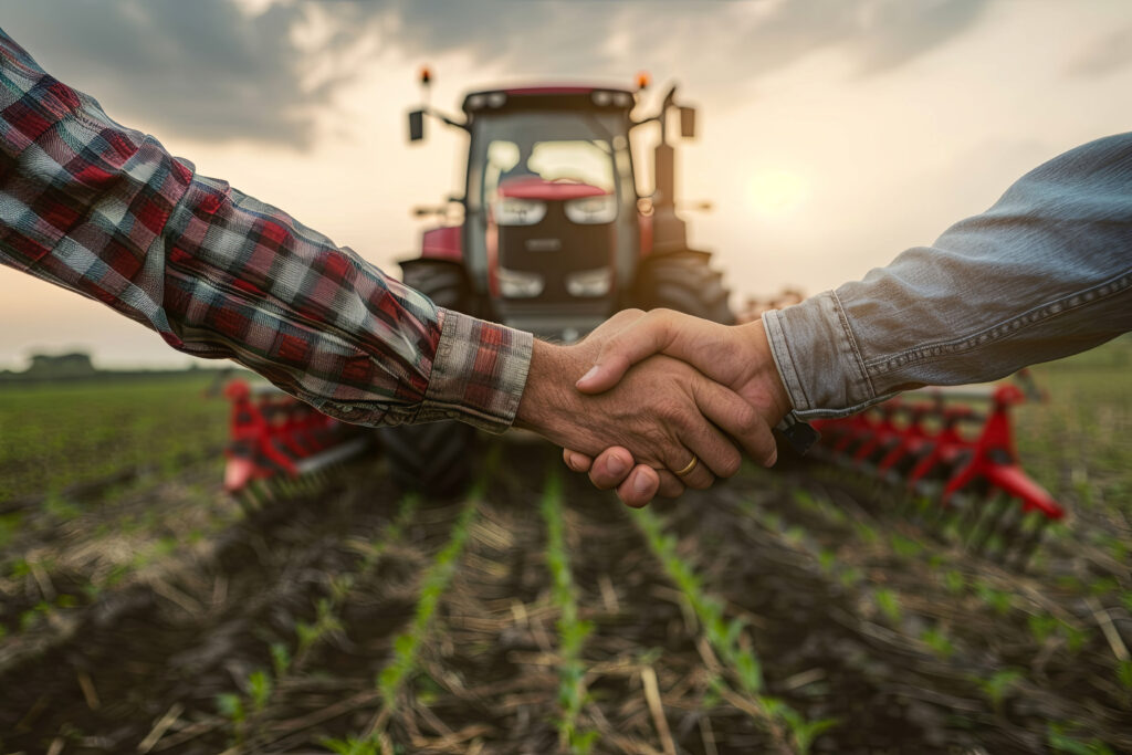 Zwei Unterarme und Hände, die sich schüttlen. Vor einem roten Traktor auf einem Feld.